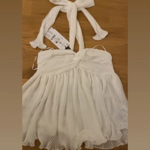 Vit omlott topp med knytning - Superfin vit topp med omlott-detalj och knyt i nacken. Klänningen har plisserad kjol och tunna axelband, vilket ger en luftig och fräsch känsla. Perfekt för sommarens alla tillfällen och lätt att matcha med accessoarer. NY PRIS 349 HELT OANVÄND PRISLAPPEN FINNS KVAR