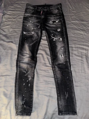 Svarta skinny jeans från Dsquared2 - Säljer ett par svarta skinny jeans från Dsquared2 med coola slitningar och färgstänk över hela byxan. Jeansen har fem fickor, låg midja och är tillverkade i Italien. Materialet är klassisk jeans med stretch för skön passform.