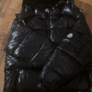 Svart Moncler pufferjacka med dragkedja - Säljer en svart pufferjacka från Moncler med glansig finish och klassisk logga på ärmen. Jackan har hög krage, dragkedja framtill och praktiska fickor. Perfekt för dig som vill ha en snygg och varm jacka med street vibe och exklusiv känsla.