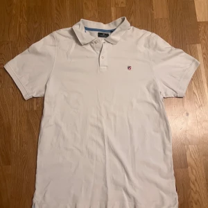 Vit pikétröja från U.S. Polo Assn. - Vit pikétröja från U.S. Polo Assn. med klassisk krage och korta ärmar. Tröjan har knappar vid halsen och en liten broderad logga på bröstet. Tillverkad i mjuk bomull som är skön mot huden. Perfekt för en clean och sportig look.