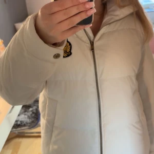 Ralph Lauren dunjacka, cream white - Säljer en vit pufferjacka från Ralph Lauren med snyggt broderat emblem i guld och marinblått på bröstet. Jackan har hög krage, men går att justera, dragkedja framtill och två sidofickor. Perfekt för dig som vill ha en clean och stilren look under vintern! 💕Org pris = 3295