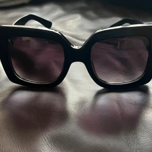 Svarta-Gucc solglasögon - Snygga solglasögon med breda, svarta bågar och stora fyrkantiga linser i mörk ton. Riktigt statement piece med retrovibe, perfekt för dig som gillar att sticka ut. Bågarna är i plast och har en cool, chunky design som ger en modern känsla.