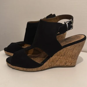 Svarta kilklackade sandaler med spänne - Snygga svarta sandaler med kilklack i kork och öppen tå. Ovandelen är i svart syntetmaterial med breda remmar och ett silverfärgat spänne vid ankeln. Sulan har blommigt mönster undertill och klacken ger extra höjd. Perfekt för dig som gillar stilren och modern look.