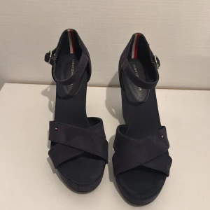 Mörkblå kilklack sandaler Tommy Hilfiger - Snygga mörkblå sandaler från Tommy Hilfiger med kilklack och plattform. Skorna har breda korslagda remmar i mocka, rund tå och justerbart spänne vid ankeln. Diskret Hilfiger-logga på remmen och klassisk röd-vit-blå detalj på insidan. Perfekta för sommarens alla tillfällen.