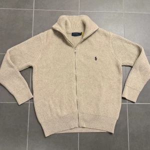 Beige ribbad kofta från Polo Ralph Lauren - Säljer en beige ribbad kofta från Polo Ralph Lauren i storlek M. Tröjan har hel dragkedja, hög krage och det klassiska broderade Polo-logot på bröstet. Materialet är mjukt och stickat, perfekt för kyliga dagar. Passformen är normal och ärmarna är långa.