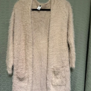 Beige fluffig kofta från Visual Clothing Project - Supermjuk och fluffig beige kofta med lång ärm och öppen framtill. Koftan har två fickor och är tillverkad i ett stickat material som ger en mysig känsla. Längd: ca 88cm, Bredd: ca 56cm. Använd fåtal gånger