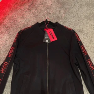 Striped Zip Hoodie från Hugo, strl S - Svart zip från Hugo med röda logoband längs ärmarna och snygg dragkedja framtill. Hoodien har ribbade muddar och är tillverkad i mjuk polyester. Perfekt för dig som gillar streetstyle och vill sticka ut med coola detaljer. Pris går att diskutera 