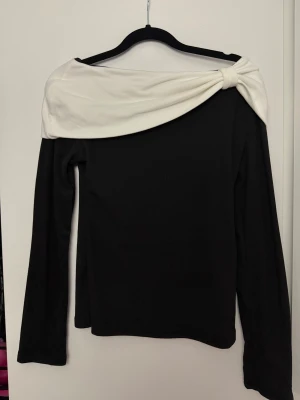 Svart topp med vit omlott-detalj - Snygg svart långärmad topp med unik vit omlott-detalj över axlarna som knyts i ena sidan. Toppen har en offshoulder-look och är perfekt för dig som vill sticka ut med en cool och stilren design.