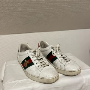 Gucci ace  - Säljer ett par vita Gucci sneakers med klassiska gröna och röda ränder på sidan samt en broderad guldfärgad bi. Skorna har snörning, rund tå och platt sula. Tillverkade i skinn med detaljer i textil. Perfekt för dig som vill ha en ikonisk och lyxig look.