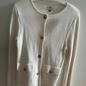 Vit kofta med knappar från Hampton Republic - Stilren vit kofta från Hampton Republic med rund halsringing och fina knappar framtill. Koftan har två fickor med lock och knappar, samt långärmad passform. Koftan är stickad i mjukt material och har en clean, klassisk look som funkar till allt