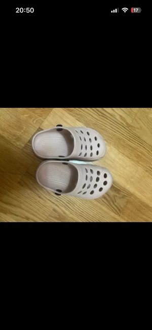 Ljusrosa sandaler med hålmönster - Säljer ett par ljusrosa sandaler med rund tå och platt sula. Skorna har ventilerande hålmönster upptill och en svart rem som kan fällas bak för extra stöd. Tillverkade i syntetmaterial, perfekta för chill dagar eller stranden.