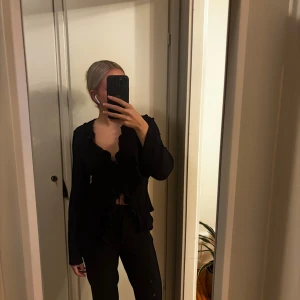 Svart blus med volang och snörning - Säljer en svart blus med långa ärmar, djup v-ringning och volangkant längs hela framsidan. Blusen har snörning framtill och är lätt croppad. Strl M