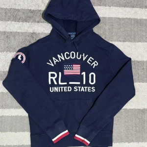 Mörkblå hoodie RL-10 Vancouver USA - Snygg och bekväm hoodie i mörkblå färg med tryck framtill: “Vancouver RL-10 United States” och en amerikansk flagga. Märke på ärmen och ribbade muddar med röd/vit detalj. Tröjan är i barnstorlek L, men motsvarar ungefär en vuxen storlek S.  Längd från axeln och ner: 57 cm Bredd under armhålorna: 43 cm  📦 Skickas snabbt & välpackat