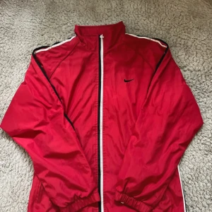 Röd Nike vindjacka med vita detaljer - Säljer en röd vindjacka från Nike med vita och svarta detaljer längs ärmarna. Jackan har hel dragkedja framtill, två sidofickor med dragkedja och klassisk Nike-logga broderad på bröstet. Perfekt för dig som gillar sportig stil. Se bilder för skick.