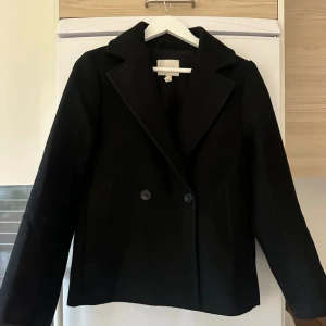 Svart skepparkavaj XS  - Snygg svart skepparkavaj i storlek XS med klassiska stora slag och dubbelknäppning framtill. Jackan har två rejäla fickor och är helsvart med stilren look. Perfekt för dig som gillar en tidlös och clean stil.