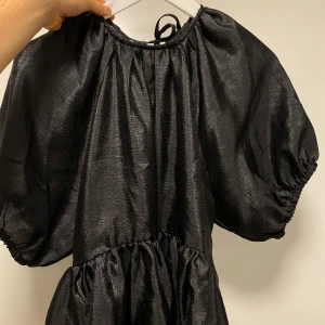 Svart festtopp med puffärm - Supersnygg svart topp med glansig finish och puffiga kortärmar. Toppen har volang nedtill och knytning i nacken samt framtill för extra detalj. 