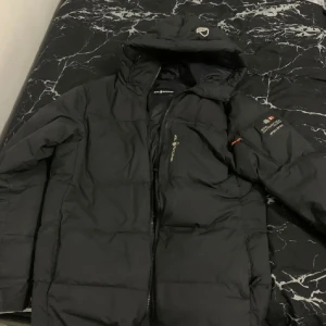 Svart dunjacka Sail Racing S - Svart dunjacka från Sail Racing med huva och dragkedja. Jackan har logga på bröstet och ärmen, samt detaljer som broderad symbol på huvan. Perfekt för kalla dagar med sin puffiga design och sportiga look.