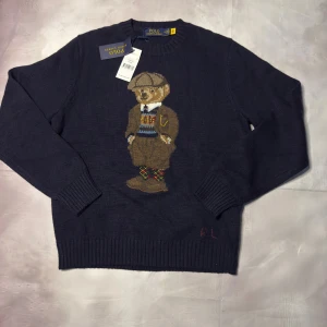 Mörkblå stickad tröja Polo Bear - Mörkblå stickad tröja från Polo Ralph Lauren med ikoniska Polo Bear-motiv på bröstet. Rund hals, ribbade muddar och RL-broderi vid nederkanten. Tröjan har lång ärm och är perfekt för dig som gillar klassisk preppy stil med en twist.