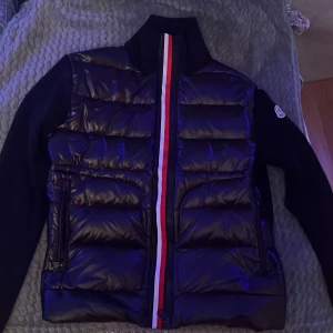 Svart jacka från Moncler med röd/vit rand - Svart jacka från Moncler med quiltad front och ikonisk röd och vit rand längs dragkedjan. Jackan har Moncler-logga på vänster ärm och två sidofickor med dragkedja. Materialet är glansig polyester och dunfyllning, med stickade svarta ärmar och krage.