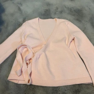 Ljusrosa omlottkofta med knytband - Superfin ljusrosa omlottkofta med långa ärmar och breda knytband i midjan. Mjuk stickad känsla och v-ringad halsringning som ger en chill och feminin vibe. Perfekt att slänga på sig när du vill ha något mysigt men ändå snyggt.