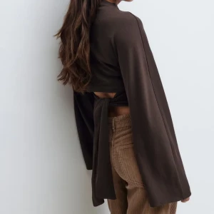 Brun croppad topp med knytband - Snygg brun croppad topp med långa ärmar och knytband i ryggen. Toppen har en enkel, stilren design med lätt hög hals och mjukt material som ger en skön känsla. Perfekt för dig som gillar trendiga detaljer och vill ha en cool look. Aldrig använd men ingen prislapp