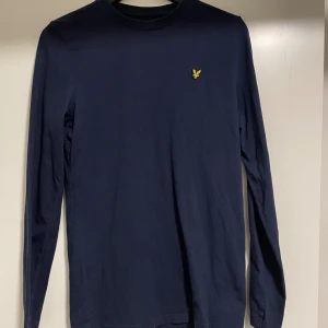 Mörkblå långärmad tröja Lyle & Scott - Snygg mörkblå långärmad tröja från Lyle & Scott med klassisk rund hals och gul broderad logga på bröstet. Tillverkad i mjuk bomull som är skön mot huden. Perfekt för dig som gillar stilrena och enkla plagg med en touch av märke. Finns 2 stycken likadana. Priset är för en tröja.