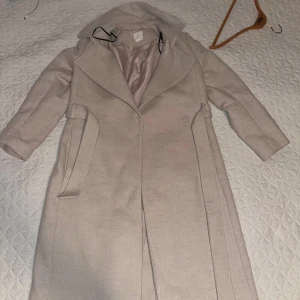 Beige kappa från H&M med bälte - Snygg beige kappa från H&M i storlek 36. Den har ett klassiskt snitt med bred krage, bälte i midjan och knäppning framtill. Fodret är i ljus satin och kappan är perfekt för höst och vinter. Stilren och enkel att matcha med olika outfits.
