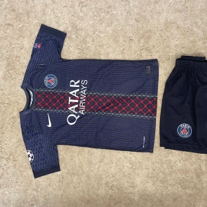 PSG Dembele #10 matchtröja Nike - Säljer en officiell Paris Saint-Germain fotbollströja med O. Dembele och nummer 10 på ryggen. Tröjan är mörkblå med röda och grå detaljer, PSG-logga på bröstet och Champions League-märke på ärmen. Materialet är lätt och andas, perfekt för träning eller match.
