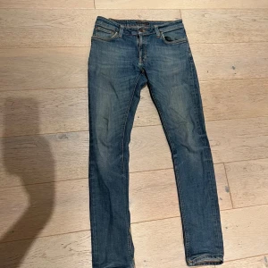 Blå slim jeans från Nudie Jeans - Snygga blå jeans från Nudie Jeans i modellen slim med klassisk femficksdesign och orangea sömmar. Jeansen har en smal passform och är tillverkade i slitstarkt denimtyg. Perfekt för dig som gillar en tajt look och vill ha ett par jeans med coola detaljer. Hål i vänster fickan