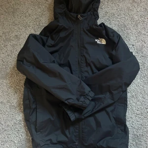 Svart vindjacka från The North Face - Svart vindjacka från The North Face med huva och dragkedja framtill. Jackan har en stilren design med logga på bröstet och ryggen, samt elastiska muddar vid ärmslut. Perfekt för blåsiga dagar