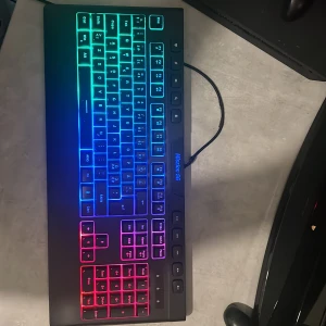 Mission SG RGB Gaming Keyboard - Snygg RGB-belyst gamingtangentbord från Mission SG med nordisk layout. Flera färglägen, mediaknappar och numeriskt tangentbord. USB-anslutning. Tangenter och belysning i fint skick, inga synliga defekter eller slitage. Perfekt för gaming och kontor.