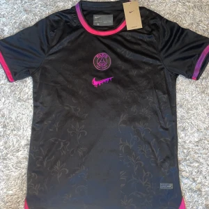 Svart PSG x Nike fotbollströja  - Snygg svart Paris Saint-Germain fotbollströja från Nike med rosa/lila detaljer och logga. Tröjan har korta ärmar, rund hals med rosa kant och subtilt blommönster i tyget. Materialet är lätt och andas, perfekt för träning eller match.
