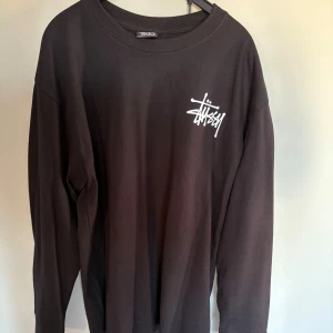 Svart långärmad Stüssy tröja - Svart långärmad tröja från Stüssy med stort vitt logotryck på ryggen och mindre logga på bröstet. Tröjan har en klassisk rund hals och är gjord i mjuk bomull som är skön mot huden. Perfekt för dig som gillar streetwear och vill sticka ut med en ikonisk look.