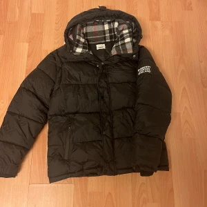 Svart Burberry pufferjacka med huva - Svart pufferjacka från Burberry med avtagbara ärmar så du kan köra väststil. Jackan har huva med rutigt foder i grått, vitt och rött. Perfekt för kalla dagar. Priset är inte huggit i sten. Hör av er vid minsta lilla fundering!