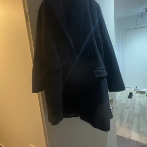 Svart kappa med klassisk look - Säljer en svart kappa med stilren design och klassisk krage. Jackan har två framfickor och knappar framtill. Perfekt för dig som gillar en enkel men snygg stil. Passar till många olika outfits och håller dig varm under kyliga dagar.