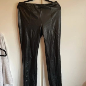 Svarta skinnbyxor med slits - Snygga svarta skinnbyxor med raka ben och slits vid benslutet. Byxorna har en clean look med sömmar framtill och är tillverkade i ett glansigt skinnmaterial som ger en cool vibe. Perfekta för dig som vill sticka ut med en edgy stil.