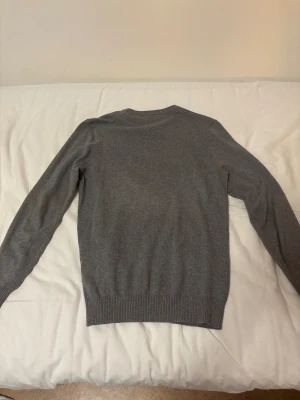 Grå stickad tröja från Lyle & Scott - Snygg grå stickad tröja från Lyle & Scott med klassisk rund hals och långärmad passform. Tröjan har märket broderad på bröstet och ribbade muddar vid ärmslut och nederkant. Perfekt för dig som gillar stilrena och enkla plagg.