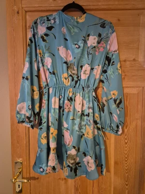 Blommig klänning från Zara - Säljer en ljusblå kort omlottklänning från Zara med v-ringning och blommigt mönster i rosa, gul och svart. Klänningen har långa ärmar med resår vid handlederna, volang nedtill och knyt i midjan för en snygg siluett. Perfekt för dig som gillar färg och mönster! Aldrig använd, prislapp kvar.