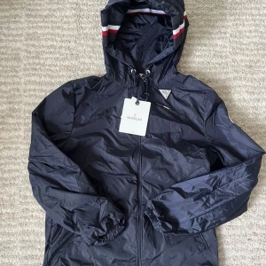 Mörkblå vindjacka från Moncler - Snygg mörkblå vindjacka från Moncler med huva och coola rödvita detaljer på kanten. Jackan har dragkedja framtill, snörning vid huvan och Moncler-logga på ärmen. Materialet är lätt och syntetiskt, perfekt för blåsiga dagar.m passar S 