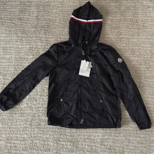 Svart vindjacka från Moncler med huva - Svart vindjacka från Moncler med huva och coola vita och röda ränder på toppen. Jackan har dragkedja, snörning vid huvan och två fickor med tryckknappar. Liten Moncler-logga på ärmen. Perfekt för dig som gillar stilren och sportig look.