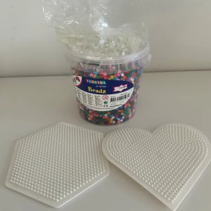 Pärlset med plattor  - Ett stort pärlset med en hink full av pärlor och två pärlplattor i form av en hexagon och ett hjärta. Perfekt för pyssel! En platta har blivit böjd när jag strykte på den, kanske går att göra platt igen? Påsen med ”genomskinliga” pärlor som är ovanpå burken lyser i mörkret!