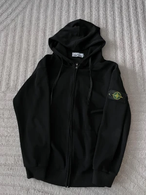 Svart hoodie från Stone Island M - Svart hoodie från Stone Island med dragkedja och klassisk patch på ärmen. Tröjan har huva med snörning och ribbade muddar. Tillverkad i mjuk bomull, perfekt för en clean och stilren look.