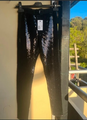 Svarta paljettleggings - Säljer ett par svarta leggings med glittriga paljetter över hela ytan. De har en elastisk midja och sitter tight mot benen, perfekta för att sticka ut med en extra glansig look. Materialet är syntet och paljett, vilket ger en cool och festlig vibe.