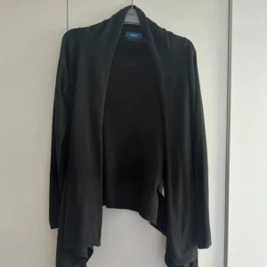 Svart omlott kofta från Mexx - Svart kofta från Mexx med långärmad design och snygg drapperad och asymmetrisk nederkant. Koftan är i mjuk material och har en loose passform som ger en chill vibe. Perfekt att slänga på sig över en T-shirt eller linne när det blir lite kyligt och som gillar stilrena och enkla plagg