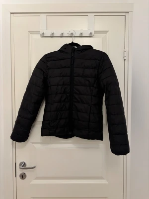 Svart pufferjacka med huva - Svart pufferjacka med huva och horisontella quiltade sömmar. Jackan har dragkedja framtill och är långärmad. Perfekt för kyliga dagar när du vill hålla dig varm och samtidigt se chill ut.