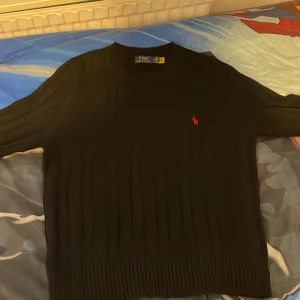 Svart kabelstickad tröja Polo Ralph Lauren - Svart kabelstickad tröja från Polo Ralph Lauren i storlek M. Klassisk design med rund hals och röd broderad logga på bröstet. Tröjan har ribbade muddar och är långärmad, perfekt för dig som gillar stilrena och tidlösa plagg.