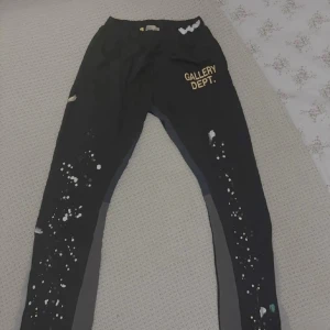 Gallery Dept. svart och grå mjukisbyxor - Säljer ett par svarta och grå mjukisbyxor från Gallery Dept. med coolt färgstänk-tryck längs benen och tryckt logga i gult på framsidan. Byxorna har elastisk midja och en relaxed passform, perfekta för en streetwear-look.