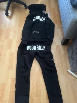 Svart Hoodrich hoodie och byxor med tryck - Svart hoodie från Hoodrich och byxor med stor vit logotyp på bröstet och detaljerat tryck på fickan och ryggen. Tröjan har huva, lång ärm och en klassisk känguruficka framtill. Materialet är mjukt och tjockt, perfekt för chill eller streetwear. Snygga vita tryck ger en cool kontrast.