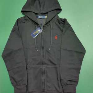 Svart hoodie från Polo Ralph Lauren - Svart hoodie från Polo Ralph Lauren med röd broderad logga på bröstet. Tröjan har huva med snörning, dragkedja framtill och två fickor. Tillverkad i mjuk bomull och har långa ärmar med ribbade muddar. Perfekt för chill dagar.