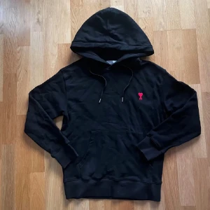 Svart hoodie från Ami Paris med rött hjärta - Svart hoodie från Ami Paris med klassisk huva och snörning. På bröstet finns ett broderat rött hjärta med bokstaven A. Tröjan har en stor magficka och ribbade muddar vid ärmslut och nederkant. Perfekt för dig som gillar stilrena streetwear-plagg.